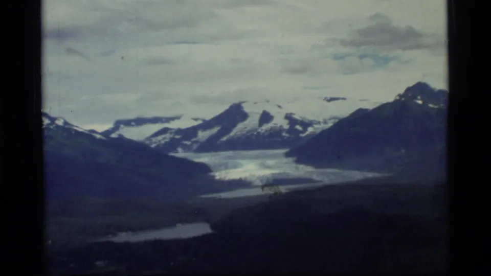 Wide 1977 8mm aerial view of Glacier Bay landscape with glacier ice, dark ridges, small lakes, and snow-capped mountains.