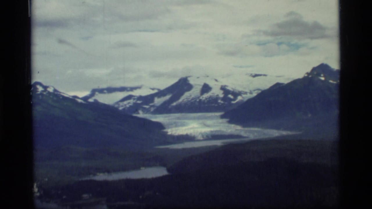 1977 8mm view over Glacier Bay, Alaska, showing snowy mountains, a glacier valley, cloudy sky, and aircraft-window edges.