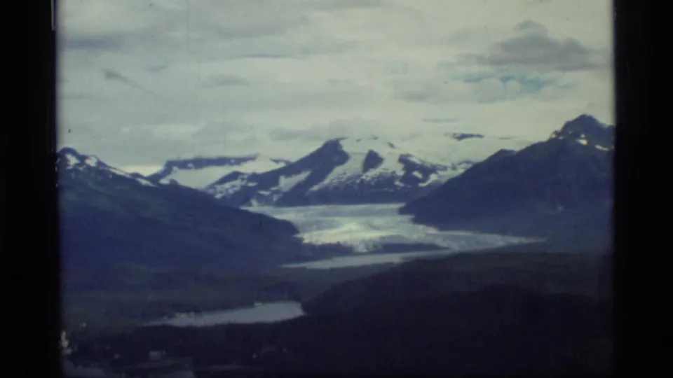 1977 8mm view over Glacier Bay, Alaska, showing snowy mountains, a glacier valley, cloudy sky, and aircraft-window edges.