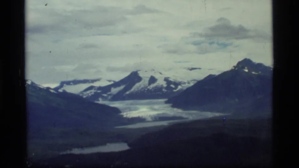 Aerial 8mm frame from Glacier Bay, Alaska, with a glacier between dark mountain slopes and snow-covered peaks beyond.