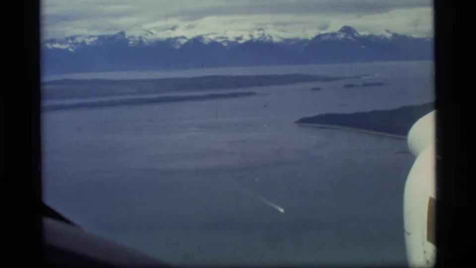 1977 Glacier Bay aerial frame showing broad water, low shoreline forms, distant snow-capped mountains, and aircraft structure at the right edge.