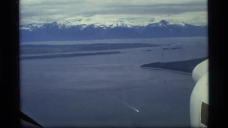 Glacier Bay, Alaska, seen from aircraft in 1977 8mm footage, with open water, distant snowy peaks, low land, and a partial aircraft edge.