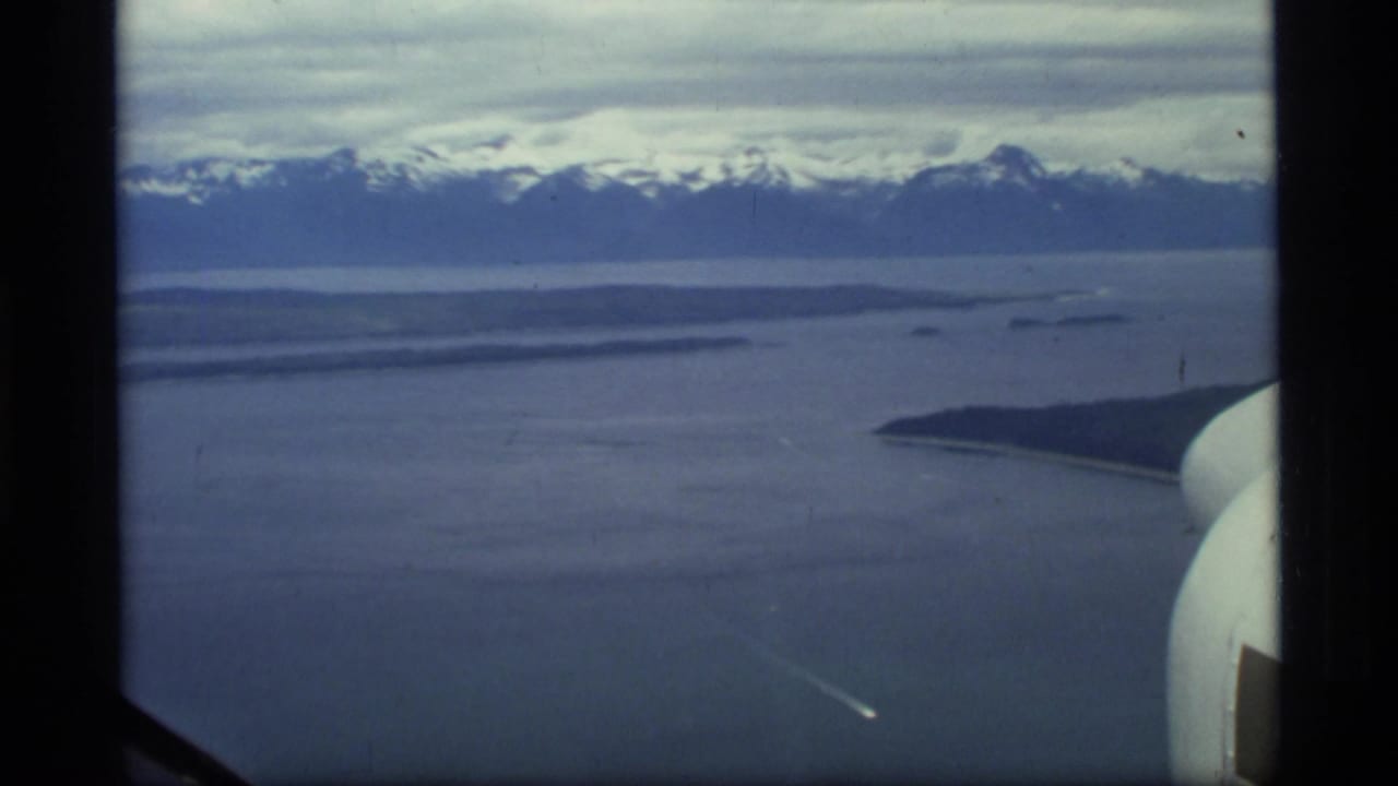Aerial view over cold water toward snow-capped mountains, with dark aircraft framing at the edge.
