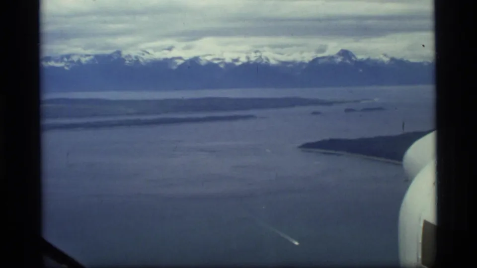 1977 8mm aerial view of Glacier Bay, Alaska, showing cold water, distant snowy mountains, and part of the aircraft window or wing frame.
