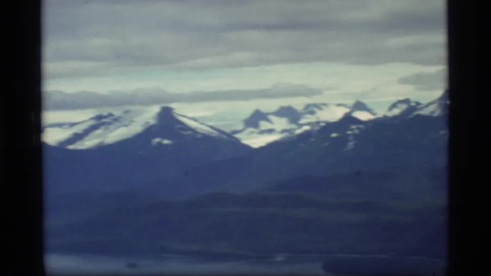 Aerial 8mm frame from Glacier Bay, Alaska, with snow-covered peaks, darker mountain slopes, cloud cover, and aircraft-window framing.