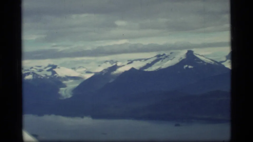 1977 Glacier Bay aerial frame showing a pale glacier between mountain slopes, snow fields, dark water, and soft 8mm color fade.