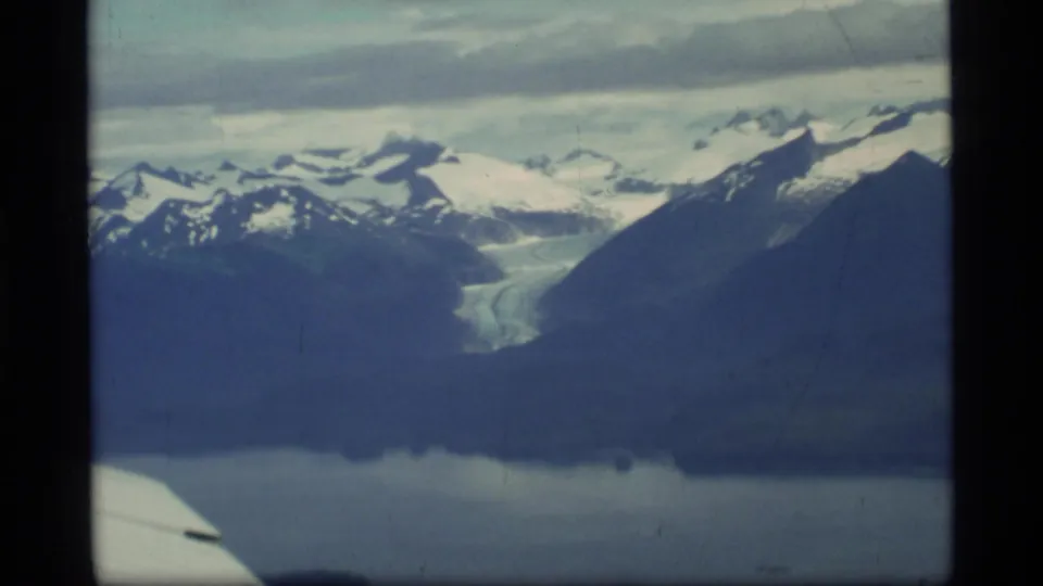 1977 Glacier Bay, Alaska aerial view with snow-covered mountains, a glacier, dark water, clouds, and part of an airplane wing in frame.