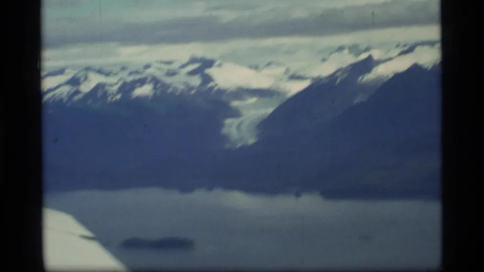 Glacier Bay aerial frame from 1977 showing a glacier between snowy ridges, dark water below, low clouds, and a partial airplane wing.