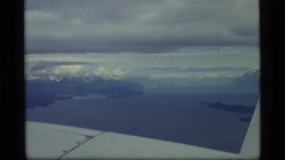1977 Alaska aerial view with the airplane wing in the foreground, dark bay water, small islands, distant snowy mountains, and heavy clouds.