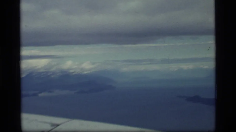 Glacier Bay, Alaska aerial frame from 1977 with a wing edge, dark water, low islands, distant snowy terrain, and layered overcast clouds.