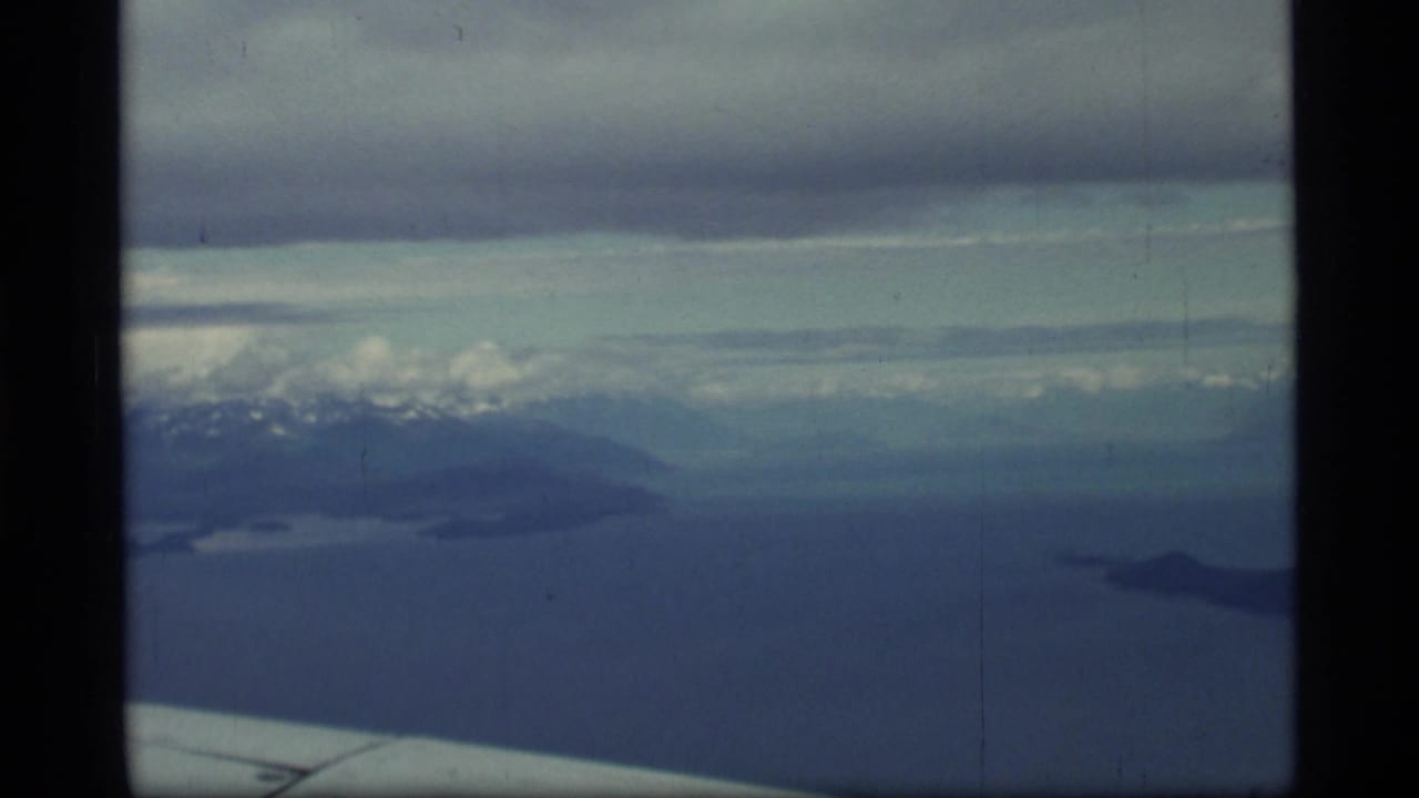 1977 Glacier Bay, Alaska airplane-window view showing blue water, distant snowy mountains, cloud cover, and part of the aircraft wing.