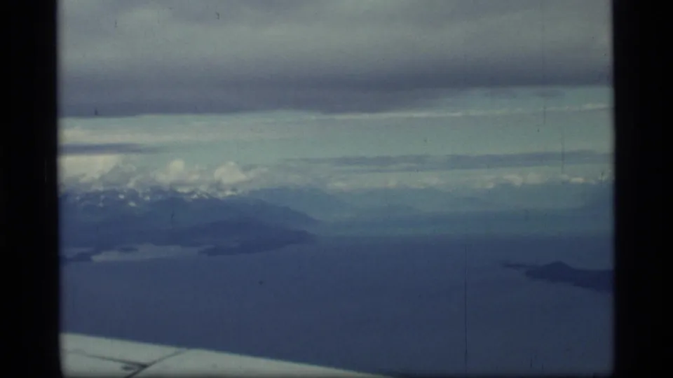 1977 Glacier Bay, Alaska airplane-window view showing blue water, distant snowy mountains, cloud cover, and part of the aircraft wing.