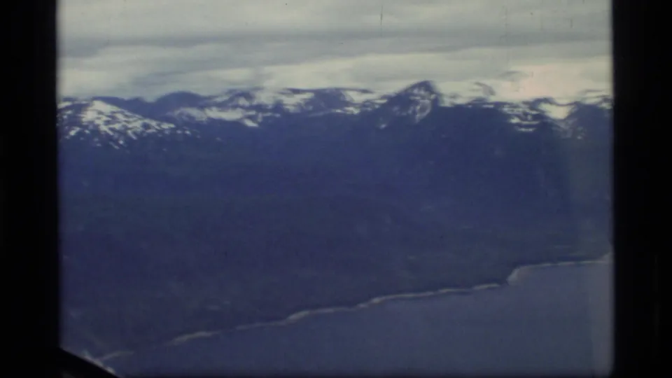 1977 Glacier Bay, Alaska aerial view with snowy mountain ridges, dark slopes, coastline, water, and airplane-window framing.