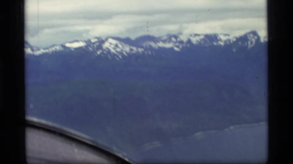 1977 Glacier Bay, Alaska view from a passenger aircraft showing dark mountain slopes, snowy ridges, water below, and window-frame edges.