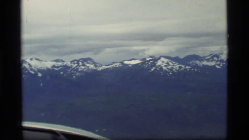 1977 Glacier Bay, Alaska aerial frame showing snow-capped mountains beneath layered clouds, dark terrain, and a small aircraft wing edge.