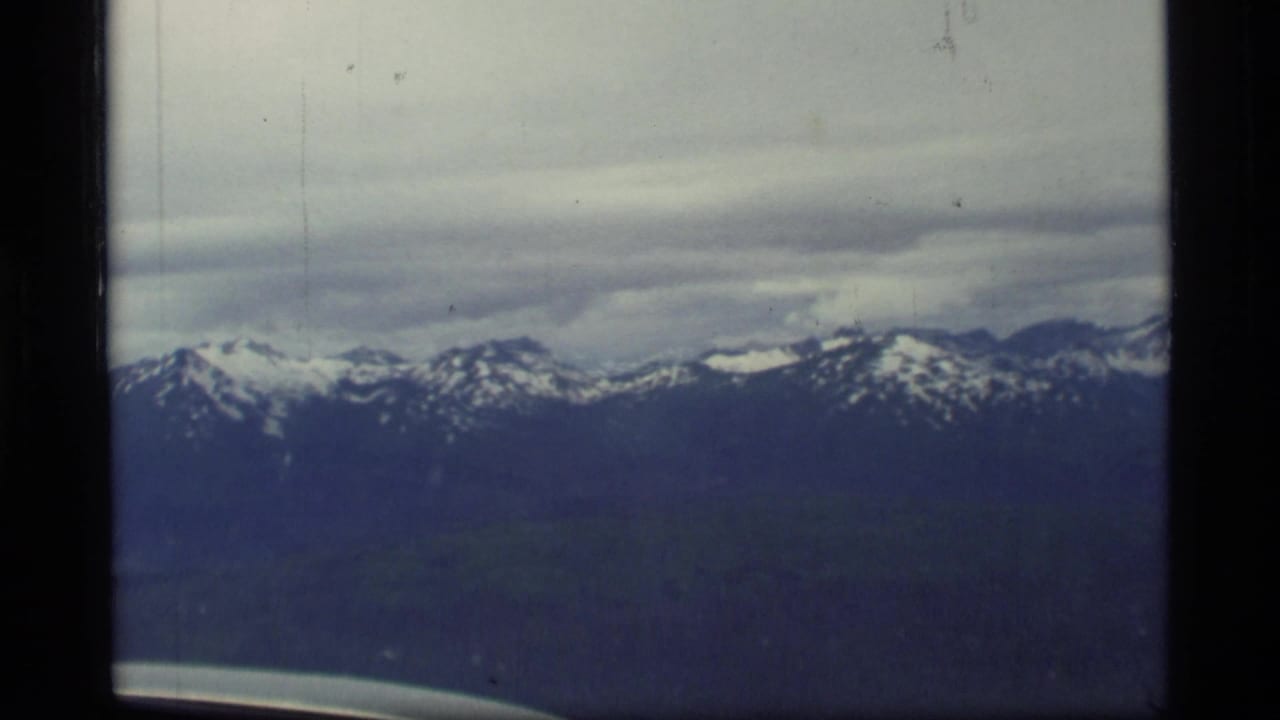1977 Glacier Bay, Alaska 8mm frame showing distant snow-capped mountains through an aircraft window with overcast sky.
