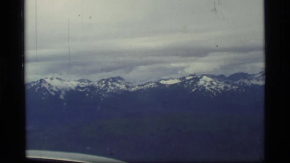 1977 Glacier Bay, Alaska 8mm frame showing distant snow-capped mountains through an aircraft window with overcast sky.