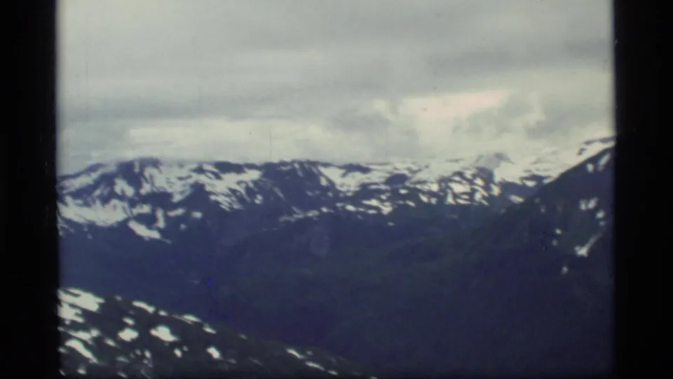 1977 home-movie frame from Glacier Bay showing a snow-streaked mountain ridge and cloudy sky from an aircraft viewpoint.