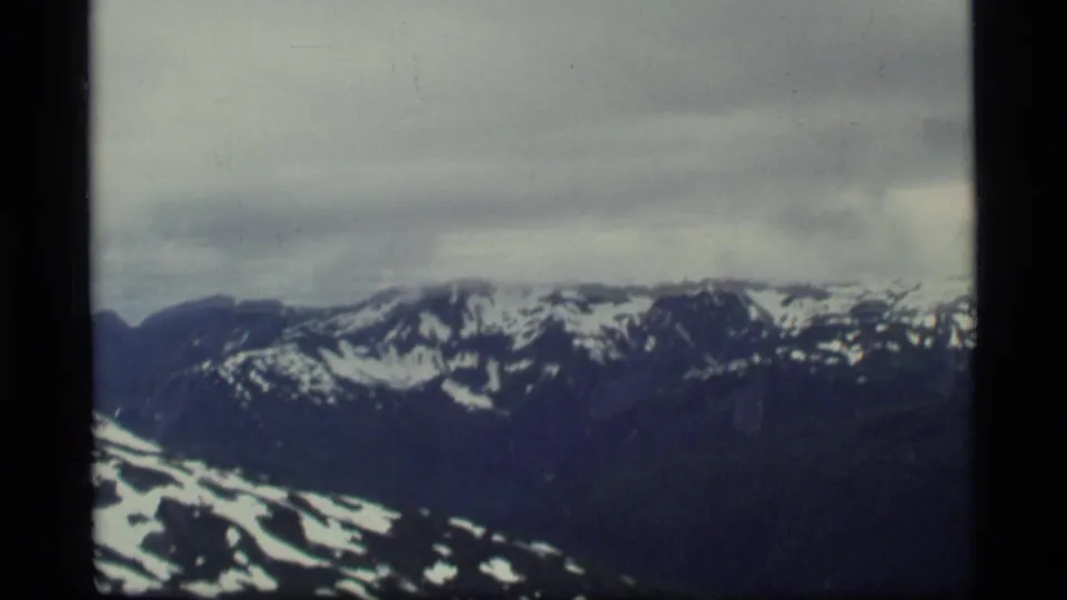 Glacier Bay, Alaska aerial frame from 1977 showing snow-capped mountains, dark slopes, and overcast sky on 8mm film.