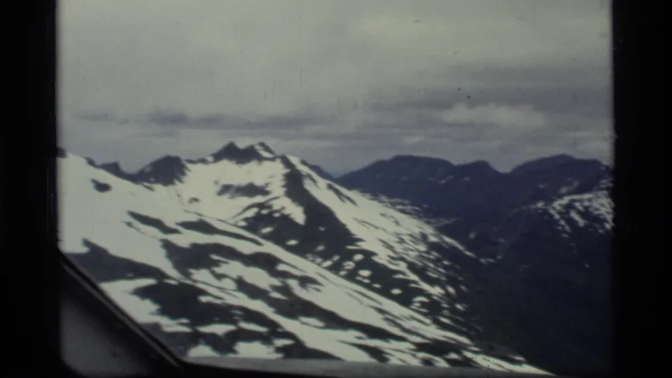 Aerial 8mm view over Glacier Bay mountain terrain, with snow patches and the airplane window frame visible in the foreground.