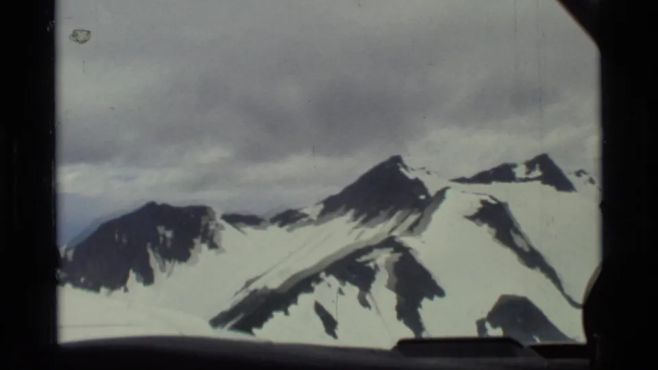 Glacier Bay aerial frame from 1977 showing jagged snowy peaks, dark rock faces, cloudy sky, and part of the aircraft window.