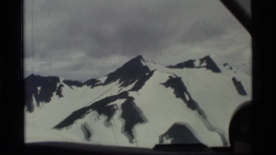 1977 8mm aerial view in Glacier Bay, Alaska, with steep snowy peaks and dark aircraft-window framing in the foreground.