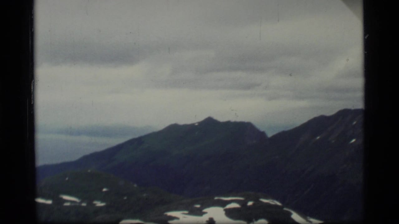 1977 8mm view from an airplane window over Glacier Bay mountains, with snow patches under a cloudy sky.