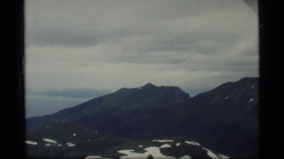 1977 8mm view from an airplane window over Glacier Bay mountains, with snow patches under a cloudy sky.