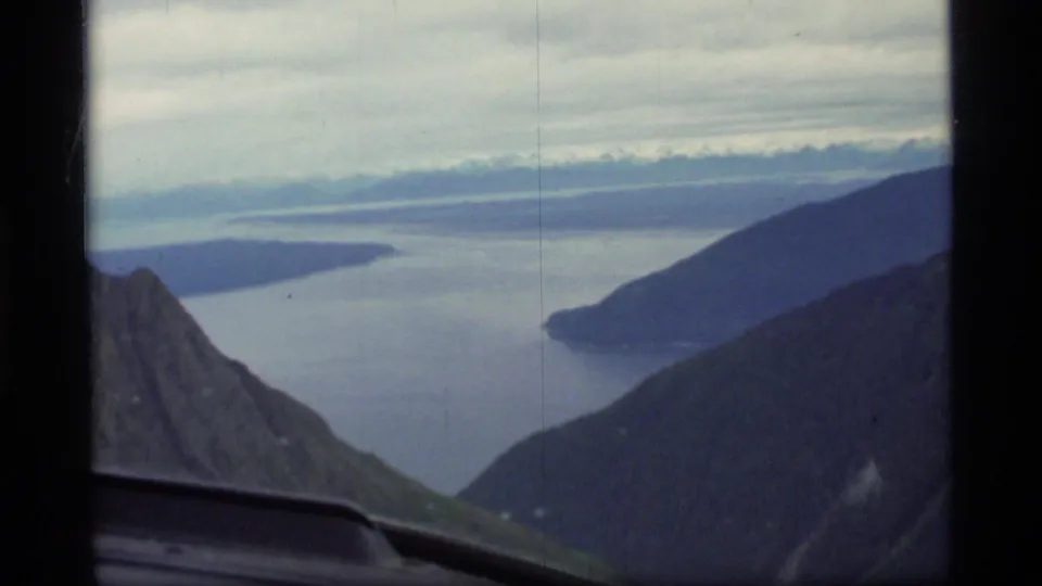 1977 Glacier Bay aerial frame with mountain slopes, a bay or inlet, distant islands, and the aircraft window edge visible.