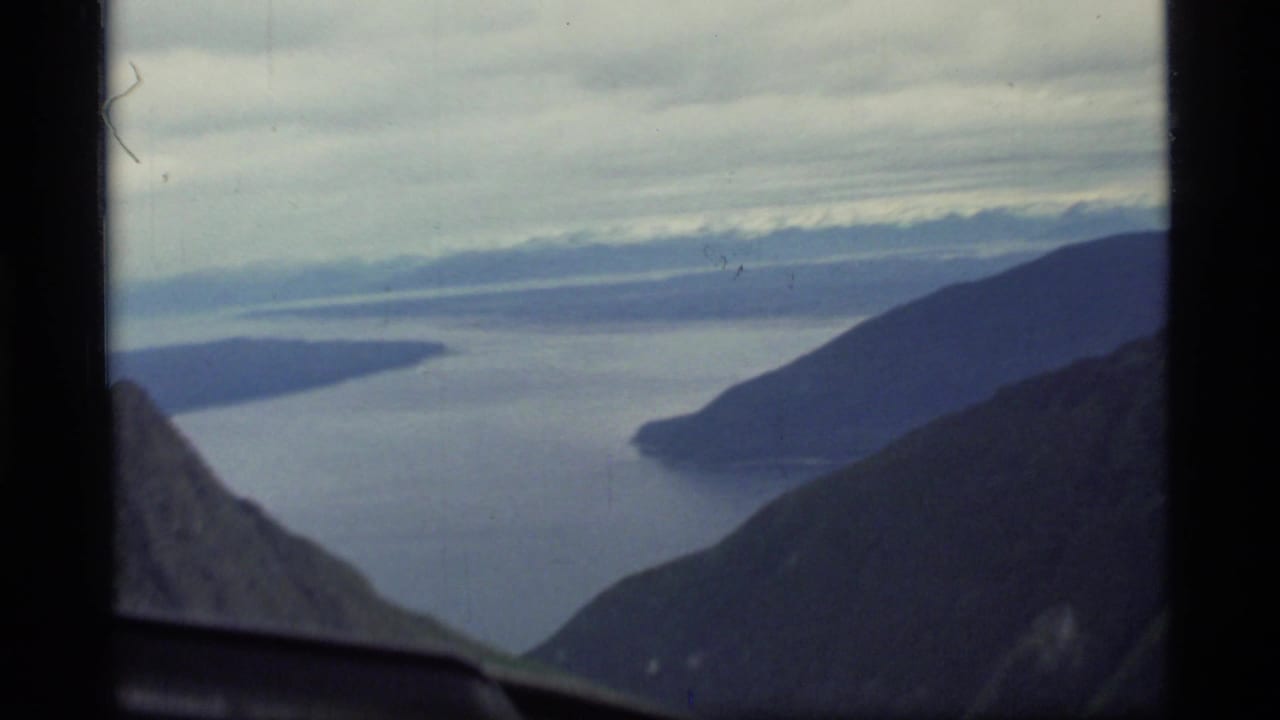 1977 8mm view over Glacier Bay, Alaska, showing water channels and dark mountain slopes through an airplane window.