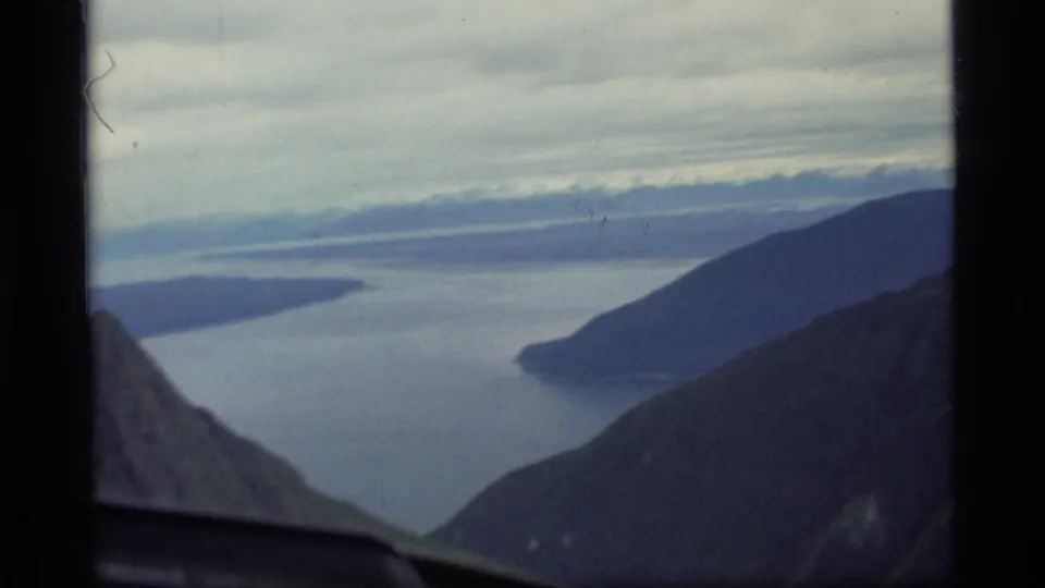 1977 8mm view over Glacier Bay, Alaska, showing water channels and dark mountain slopes through an airplane window.