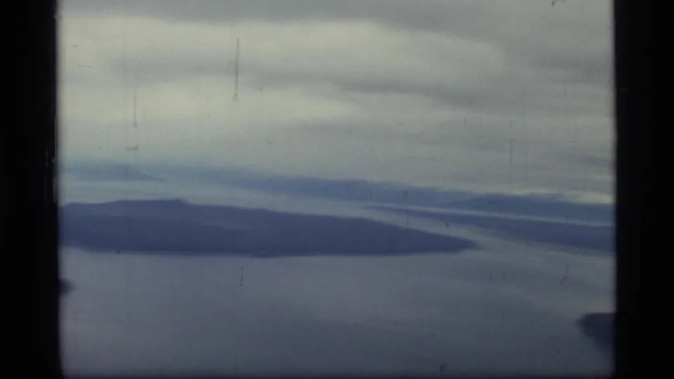 Aerial 8mm frame from Glacier Bay, Alaska, with an island-like landform, surrounding water, and overcast sky.