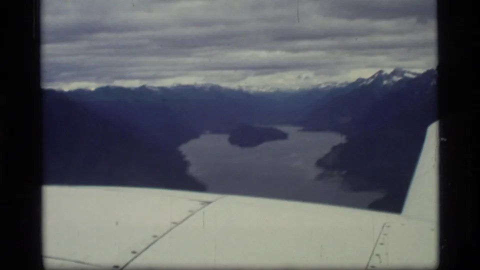 1977 Glacier Bay aerial frame with the airplane wing visible beneath dark mountains, water, islands, and snow-touched peaks.