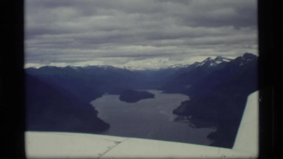 1977 8mm aerial view over Glacier Bay, Alaska, showing winding water, mountain ridges, clouds, and part of an airplane wing.