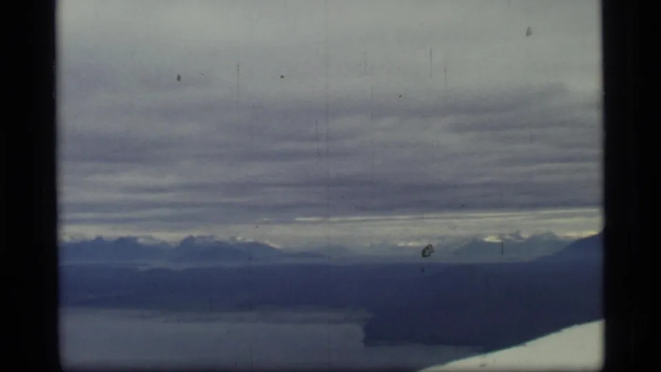 Aerial 8mm frame from Glacier Bay, Alaska, with layered clouds, distant ridges, water, and the airplane wing visible at the edge.