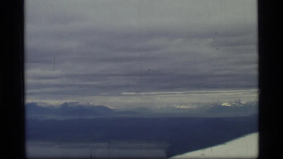 1977 Glacier Bay aerial frame showing a low cloudy sky, distant mountain silhouettes, water, and the airplane wing along the bottom edge.