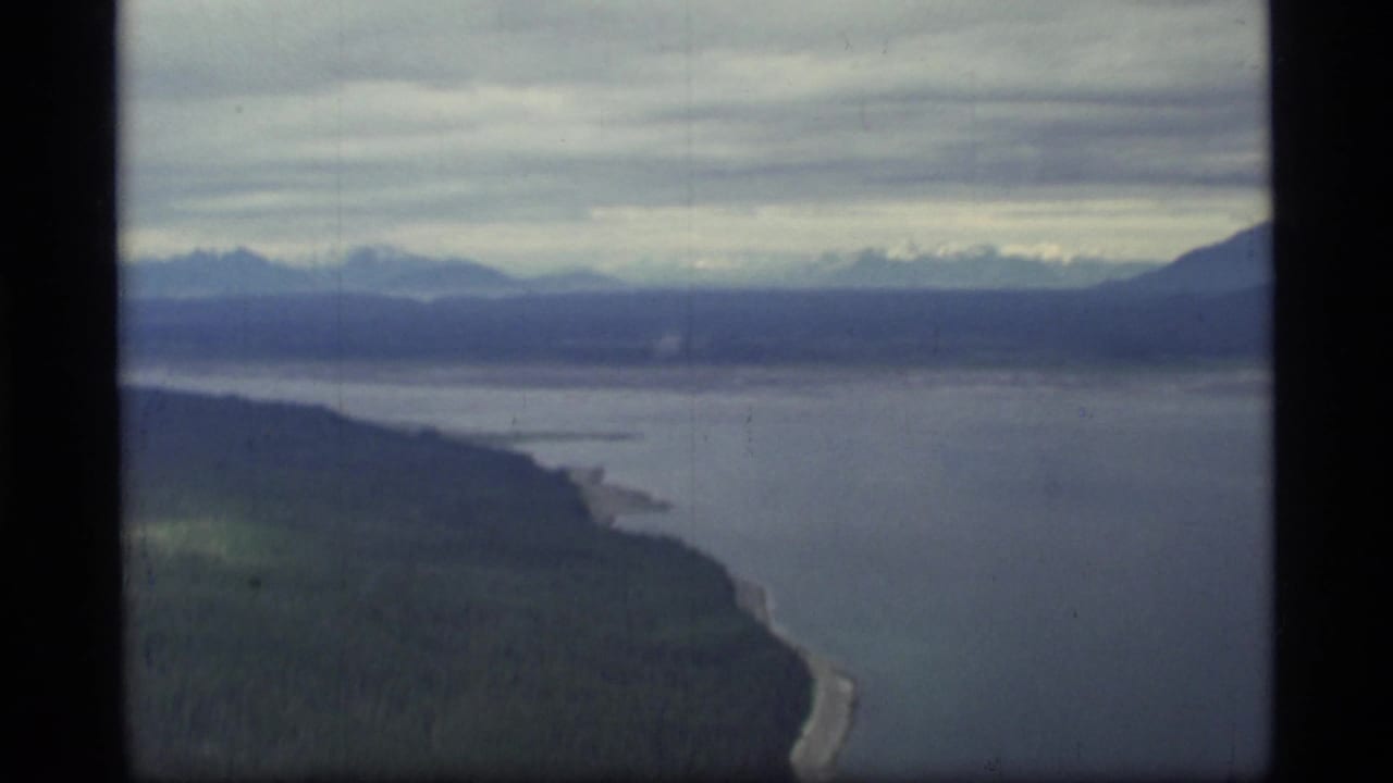 1977 8mm aerial view over Glacier Bay, Alaska, showing forested shore, calm coastal water, distant mountains, and overcast sky.