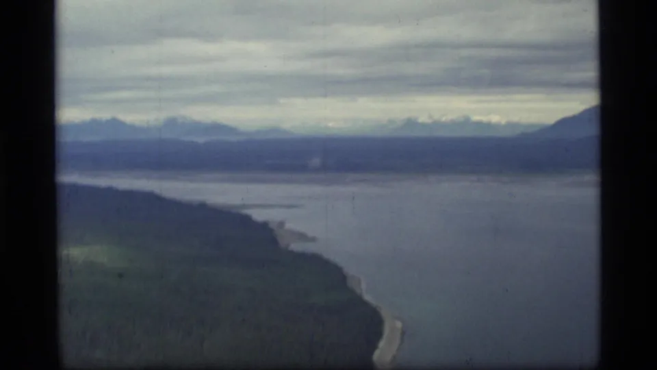 1977 8mm aerial view over Glacier Bay, Alaska, showing forested shore, calm coastal water, distant mountains, and overcast sky.