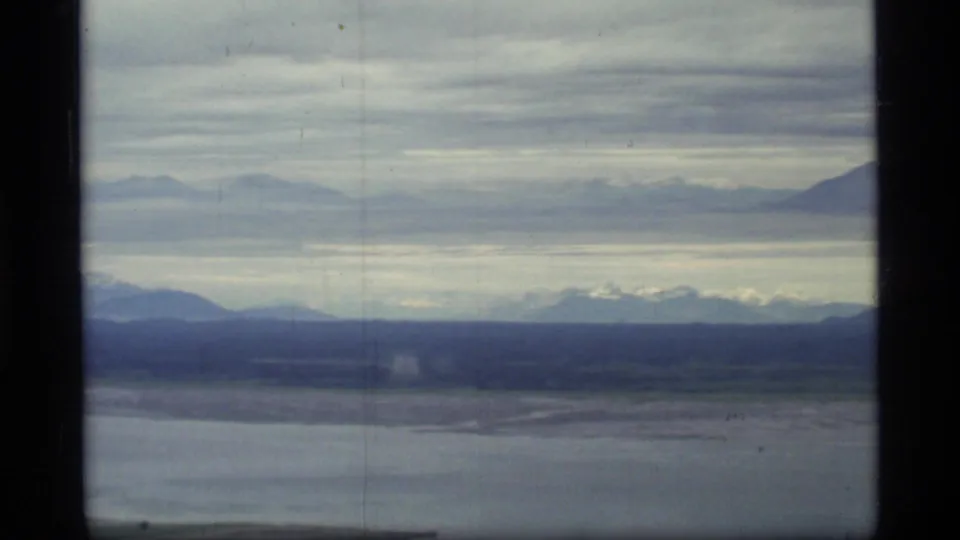 1977 Glacier Bay aerial frame with broad water, dark shoreline, snow-touched distant mountains, and a low overcast cloud deck.