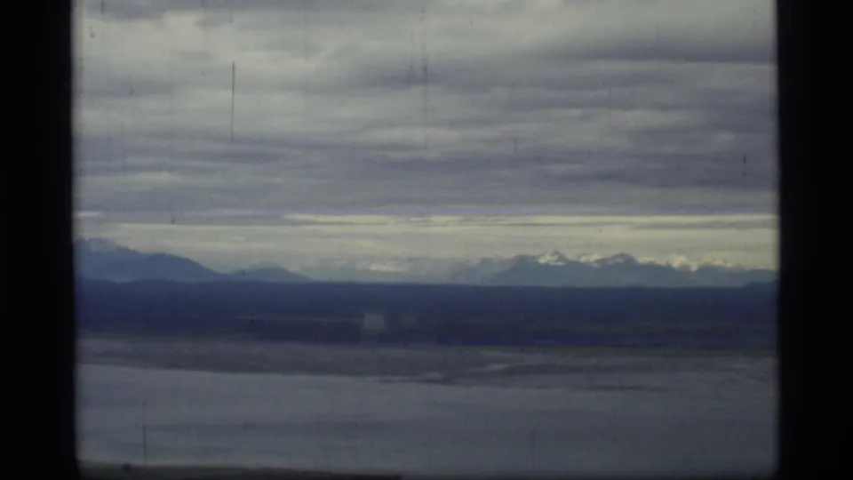 8mm aerial view from Glacier Bay, Alaska, with gray water, dark landforms, distant snowy peaks, and visible home-movie film wear.