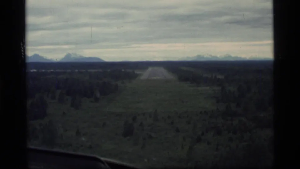 1977 8mm aerial view over Glacier Bay area forest, showing a straight cleared corridor, dark tree cover, distant mountains, and overcast sky.