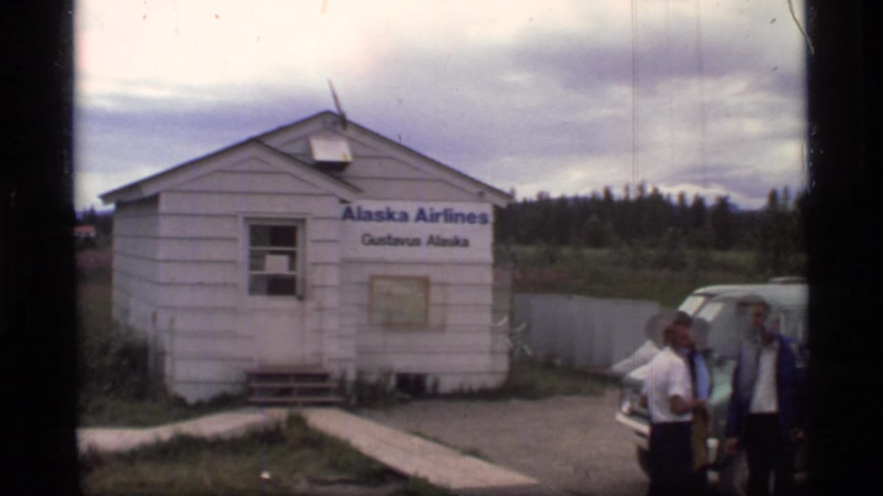 White Alaska Airlines terminal building in Gustavus, Alaska, with a few visible passengers beside a parked car.