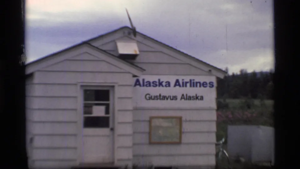 Closer 1977 view of the white Alaska Airlines Gustavus Alaska terminal with window, wall sign, and framed notice below.