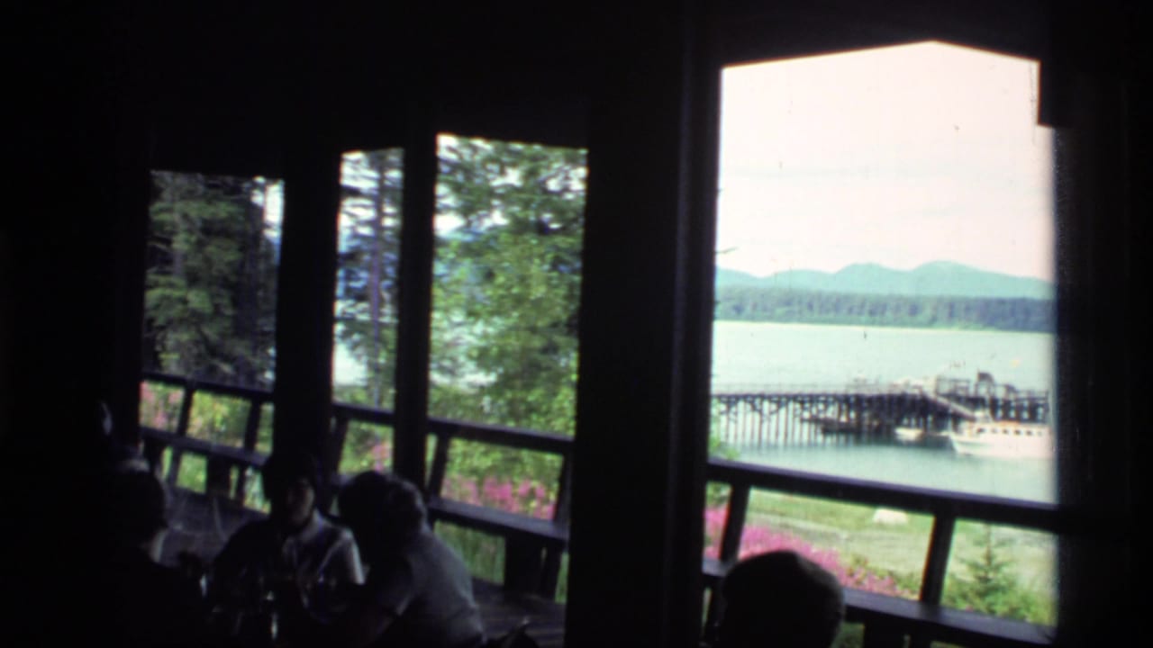 Visitors sit in silhouette by large windows overlooking a lake, pier, boat, trees, and mountains.