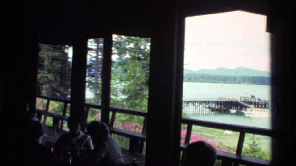 1977 Glacier Bay home-movie frame with silhouetted visitors inside a lodge facing windows over a lake, pier, small boat, evergreens, and mountains.