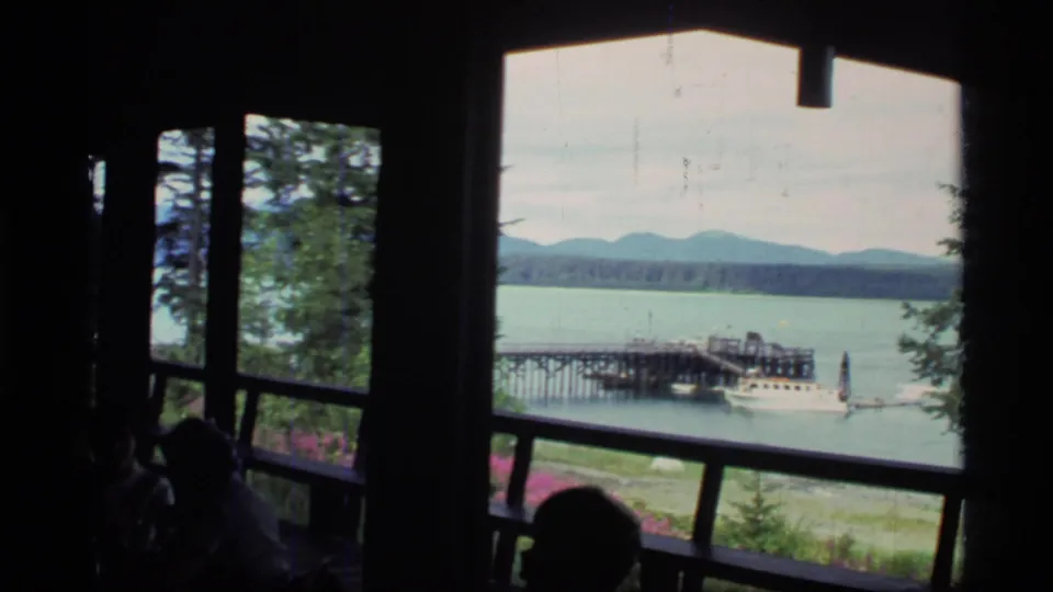 Glacier Bay lodge interior in 1977, with visitors in shadow beside windows looking out at a lake, docked boat, trees, and mountain shoreline.