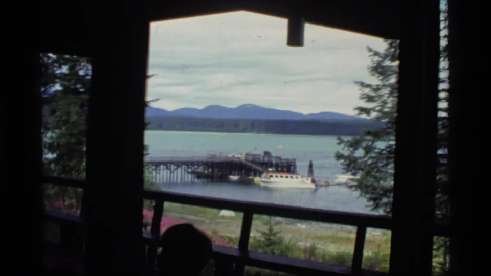A 1977 Glacier Bay 8mm frame looks past a lodge railing and window toward a calm lake, wooden pier, small boat, evergreens, and distant mountains.