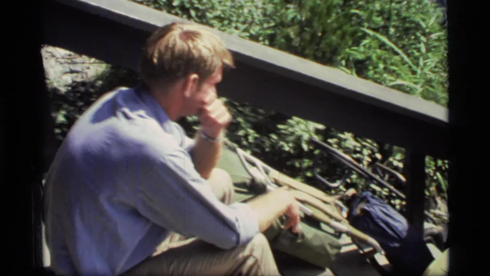 A visitor in casual 1970s clothing sits near a lodge railing in Glacier Bay, with bags beside him and vegetation beyond the deck.