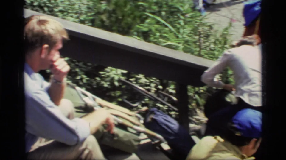 1977 Glacier Bay home-movie frame showing hikers resting beside a wooden railing with backpacks and blue caps visible.