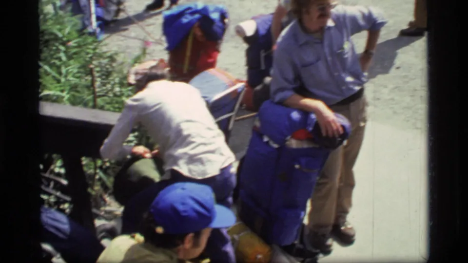 1977 Glacier Bay footage shows hikers gathered on a path, with blue packs and caps prominent in the group.